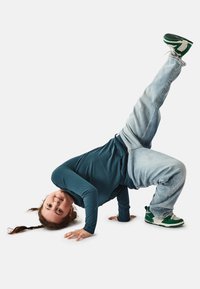 Breakdancing child in teal long-sleeve shirt, light blue jeans, and green and white sneakers, balancing on one arm while upside down.