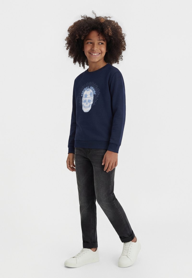 Junge mit lockigem Haar, der einen marineblauen Pullover mit Totenkopf- und Fußballmotiv, schwarze Jeans und weiße Turnschuhe trägt, steht und lächelt.