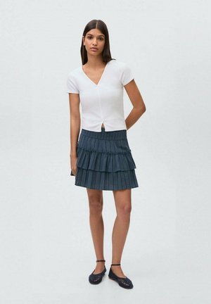 Top bianco a maniche corte con scollatura a V e dettaglio cut-out, abbinato a una gonna a strati blu scuro. Indossato con scarpe piatte blu navy.