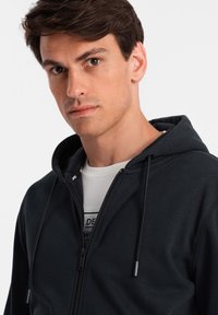 Sudadera con cremallera oscura hecha de tela suave, que cuenta con una capucha con cordón, cremallera frontal y un diseño liso sin patrones o adornos visibles.