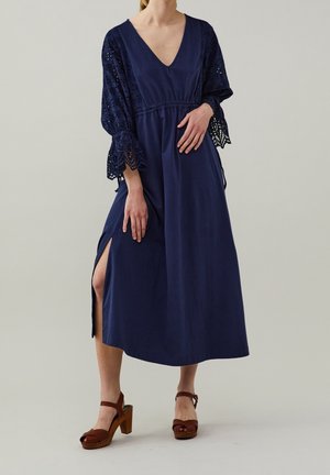 Robe bleu marine avec décolleté en V, manches longues détaillées en dentelle et fente sur le côté. Portée avec des sandales à plateforme brunes avec une large bride.