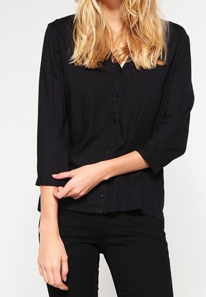 Frau mit blondem Haar trägt ein langärmliges schwarzes Hemd und schwarze Hose und posiert mit einer Hand, die den anderen Arm berührt.