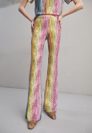 Henkilö, joka pukeutunut korkeavyötäröisiin, monivärisiin siksak-kuvioisiin liehupöksyihin ja vastaavaan yläosaan, seisoo käsivarret lanteilla ja ruskeissa kärjellisissä kengissä.