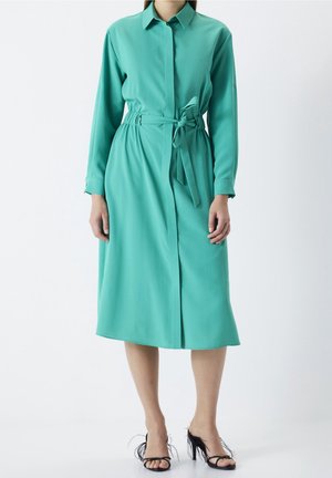 Femme portant une robe-chemise ceinturée à manches longues couleur sarcelle et des talons ouverts noirs ornés de plumes, debout devant un fond uni.