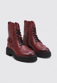 Botas de tobillo de imitación cuero rojo con un acabado texturizado, cordones negros, puntera redondeada, suela gruesa negra y detalle de correa lateral.