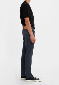 Jeans de mezclilla azul oscuro con corte recto, que presentan un parche de cuero marrón en la cintura y zapatillas de lona negras.
