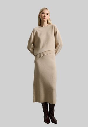 Conjunto de suéter de punto beige y falda midi con hombros caídos y dobladillos acanalados, combinado con botas marrones brillantes hasta la rodilla.