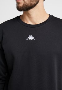 Man i svart tröja med rundad halsringning och en liten vit broderad logotyp av två sittande figurer ryggen mot ryggen på bröstet.