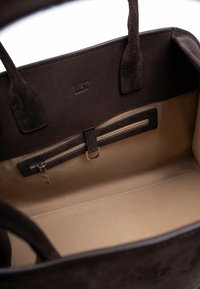 Interno di una borsa a mano in camoscio marrone con rivestimento beige, tasca con cerniera e scomparti organizzati. Logo discreto impresso all'interno.