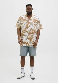 Chemise marron à manches courtes avec imprimé de feuilles blanches, assortie à un short en denim bleu clair. Le modèle porte des baskets blanches et des chaussettes mi-mollet.