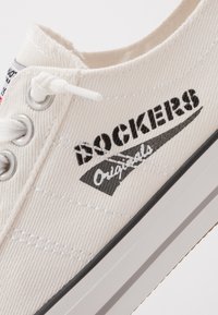 Canvas-Sneaker in Off-White mit schwarzem "Dockers Originals"-Logo, runder Zehenbereich, Schnürverschluss und Gummisohle mit beigen Akzenten.