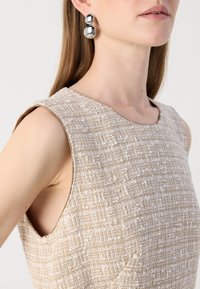Femme portant un haut beige texturé sans manches et de grandes boucles d'oreilles argentées à double boule avec des cheveux bruns raides.