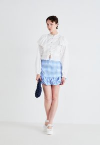Hvit, volangbluse med blå rutete miniskjørt som har volangkant. Modellen holder en mørkeblå strukturert clutch og har på seg hvite platåsko.