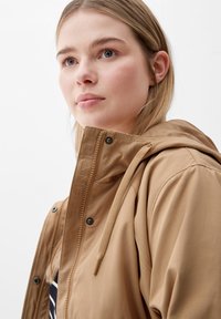 Braune wasserdichte Jacke mit Kapuze, ausgestattet mit einem strukturierten Kragen und gesticktem "TRIANGLE"-Logo, verschlossen mit Druckknöpfen und einem Reißverschluss.