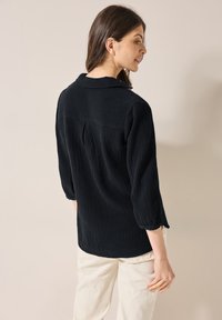 Camicia nera con texture, colletto e maniche a tre quarti, caratterizzata da una piega sul retro. Abbinata a pantaloni chiari.