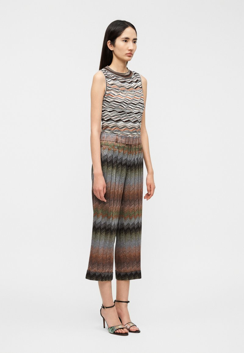Knitted sleeveless top with wave pattern in black, cream, and rust, paired with wide-leg, multicolored zigzag patterned pants and heels.