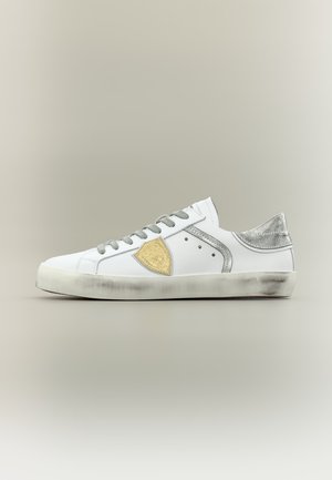 Λευκό sneaker χαμηλής κοπής με γκρι κορδόνια, μεταλλική ασημένια φτέρνα και χρυσό εμβληματικό ασπίδα στο πλάι με ελαφρώς φθαρμένη σόλα.
