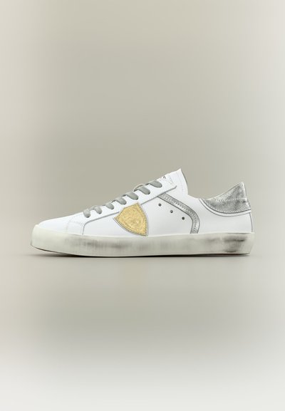 Λευκό sneaker χαμηλής κοπής με γκρι κορδόνια, μεταλλική ασημένια φτέρνα και χρυσό εμβληματικό ασπίδα στο πλάι με ελαφρώς φθαρμένη σόλα.
