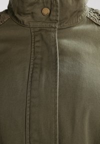 Olivgrön jacka med hög krage, knappdetaljer och texturerade virkade accenter på axlarna. Tillverkad av slitstarkt tyg.