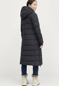 Lang sort pufferjakke med hætte, med vertikale kanaler og en lynlåslomme på siden. Bar over blå jeans og hvide støvler.