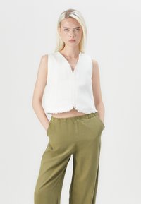 Witte, cropped, mouwloze top met rafelige randen, gecombineerd met olijfgroen wijde broek met elastische tailleband. Effen achtergrond.