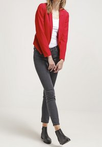 Blazer rojo ajustado con mangas largas, combinado con una blusa blanca y jeans skinny gris oscuro. Botines negros completan el conjunto.