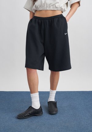 Personne portant un short noir Nike longueur genoux, des chaussettes blanches et des chaussures de sport noires sur un tapis bleu, les mains dans les poches.