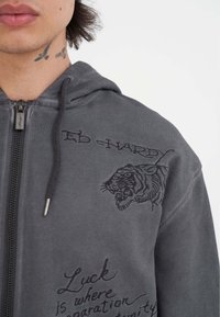 Sudadera gris con cremallera y gráfico de un tigre con texto bordado. Presenta una capucha con cordón y hardware de cremallera plateado.
