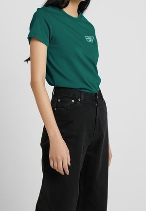 Kobieta w turkusowej koszulce z logo Vans wsuniętej w wysokie czarne jeansy, stojąca na tle jednolitego jasnego tła.