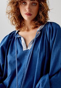 Blusa in denim di un blu intenso con scollatura a V, bordo in pizzo bianco e maniche arricciate. Il tessuto è morbido, ha una vestibilità comoda e si annoda alla scollatura.