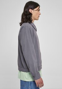 Chaqueta ligera gris con cuello, bolsillos laterales y puños elásticos, llevada sobre una camiseta verde y jeans azules.