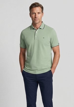 Man draagt lichtgroen poloshirt met korte mouwen en donkerblauwe broek, staat met handen in zakken, neutrale achtergrond.
