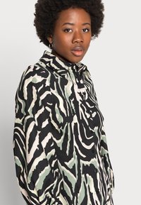 Camisa preta e creme de botões com padrão abstrato verde, apresentando um colarinho pontudo e mangas longas. Tecido texturizado com corte descontraído.