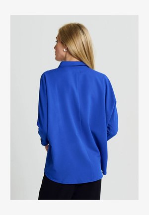 Saténová modrá blúzka s golierom, dlhými rukávmi a voľným strihom. Obsahuje zadný sklad a zaoblený lem, skombinovaná s čiernymi nohavicami.