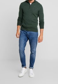 Mörkgrön zip-up tröja med ribbade muddar och fåll, tillsammans med blå slim-fit jeans och vita sneakers. Enkel, avslappnad outfit.