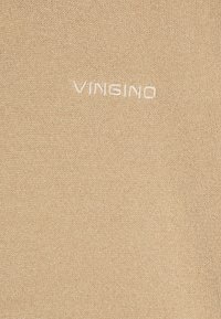 Beige gestrickter Stoff mit dem geprägten Text "VINGINO" in hellem Garn, der eine glatte Textur und eine einheitliche Farbe aufweist.