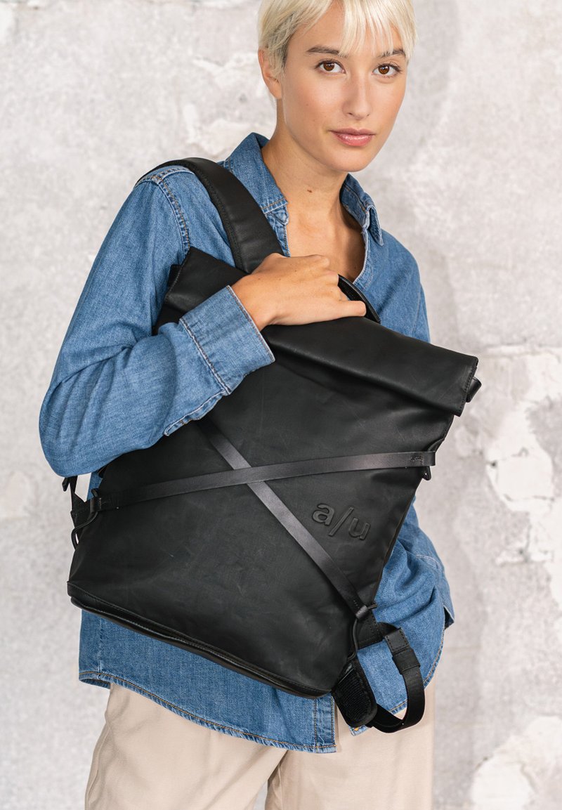 Junge Person mit kurzen blonden Haaren trägt ein blaues Jeanshemd und beige Hosen, hält eine große schwarze Leder-Umhängetasche.