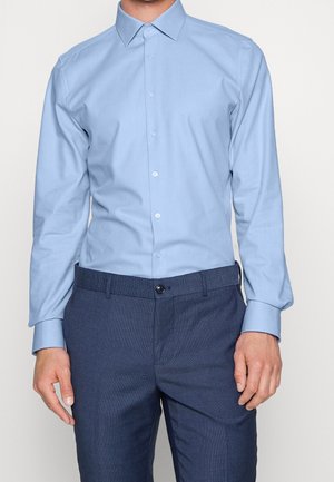 Mężczyzna ubrany w jasnoniebieską koszulę na guziki z długim rękawem, włożoną do ciemnoniebieskich eleganckich spodni ze skórzanym czarnym paskiem.