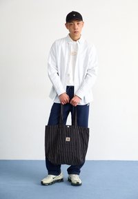 Bolso tote de rayas negras con logo, llevado con una camisa de rayas blancas, una camiseta blanca lisa y pantalones azul marino. Zapatillas deportivas en tonos neutros.