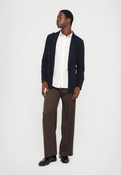 Double-breasted navy blazer over a white striped shirt, paired with brown wide-leg trousers and black shoes. Minimalist design, tailored fit.