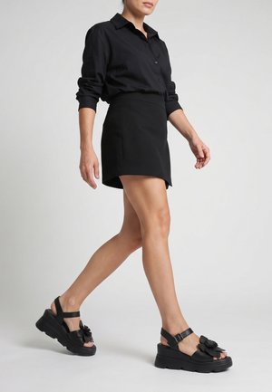 Femme marchant, portant un haut à manches longues noir, une mini-jupe noire et des sandales plateformes noires avec des décorations florales.