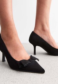 Svarta högklackade skor med spetsig tå i mjukt mockaimitation och en rosett på framdelen. Stilletthäl. Synlig ankel och vaden.