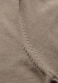 Ruskea kevyt kangas, jossa on sileä rakenne, ja sivusaumassa siistit, pienet tikit. Lähikuva korostaa ompelun yksityiskohtia.