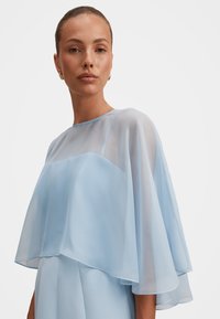 Γυναίκα που φοράει ένα διαφανές, φωτεινό μπλε, αέρινο κάπα πάνω από ένα strapless φόρεμα, με τα μαλλιά τραβηγμένα πίσω και μικρά χρυσά σκουλαρίκια κρίκους.