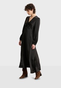 Vestido maxi de satén negro con largas mangas abullonadas, escote en V profundo, detalle de lazo en el pecho y abertura lateral. Combinado con botas de tacón marrón.