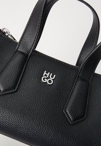 Black textured leather handbag with dual handles, silver zip, and a metal logo embossed on the front. Features angular design accents.