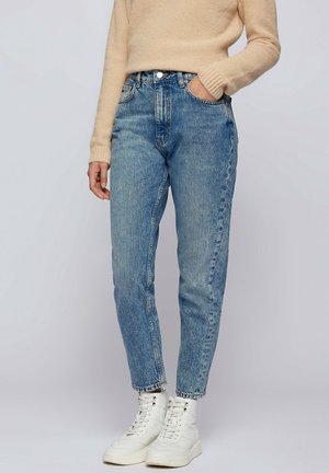 Osoba ubrana w jasnoniebieskie dżinsy z wysokim stanem, beżowy sweter oraz białe sznurowane botki za kostkę, stojąca z jedną ręką w kieszeni.