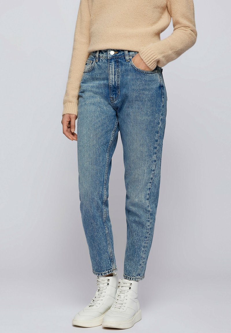 Personne portant un jean taille haute bleu clair, un pull beige et des bottines blanches à lacets, debout avec une main dans la poche.