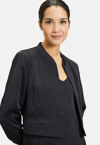 Zwarte blazer van gladde stof met een cropped ontwerp, opstaande kraag en open voorkant. Gestyleerd over een donkere top met lange mouwen.
