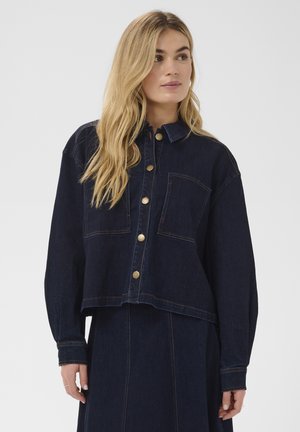 Bleg blondine iført mørkeblå denimjakke med guldknapper og matchende denimnederdel, der kigger let til siden mod en ensfarvet baggrund.
